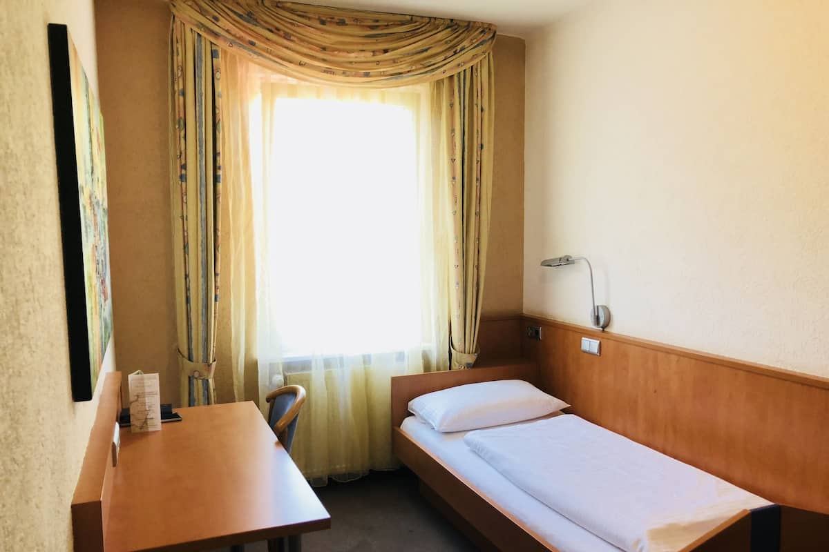 Ein kleines Hotelzimmer mit einem Einzelbett, einem Holztisch, einem Stuhl, einem Fenster mit Vorhängen und einer Wandlampe.
