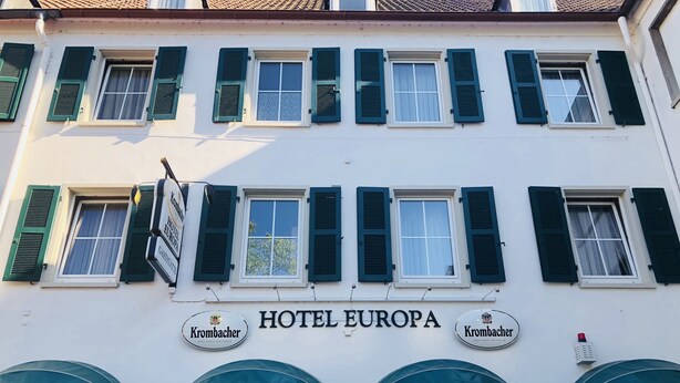 Un bâtiment doté de nombreuses fenêtres, de volets verts et d’une enseigne indiquant « HÔTEL EUROPA ».