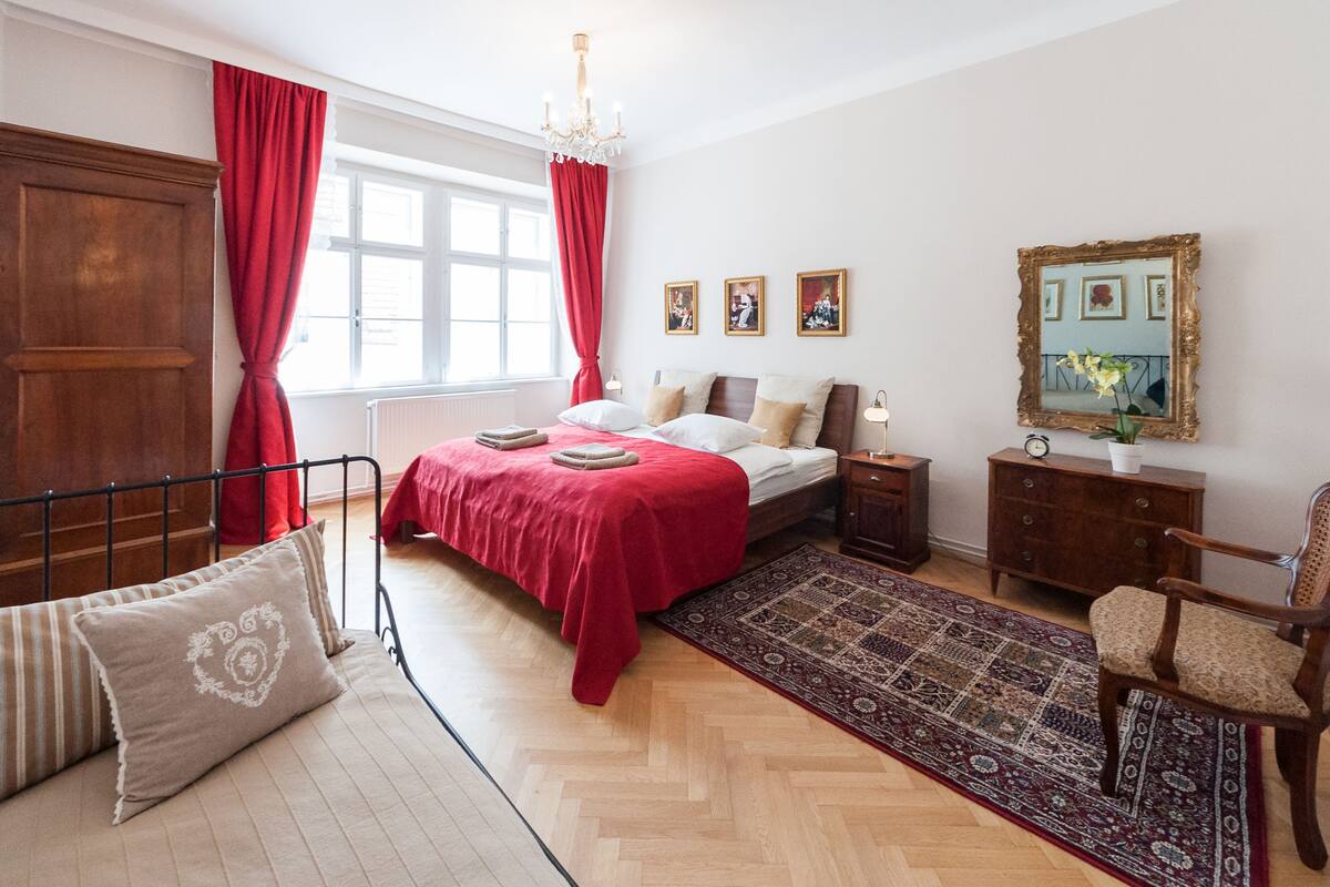 A bedroom with a bed, a wooden dresser, a chair, a nightstand, a chandelier, and a window with curtains.