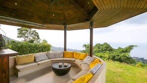 Un espace extérieur aménagé, abrité et doté d’un canapé incurvé et d’une table basse centrale, le tout entouré d’une végétation luxuriante et avec des collines en arrière-plan.