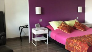 A modern bedroom with a purple accent wall, a bed with colorful bedding, a white bedside table with a clock, and a small desk with a chair. - La Ferme du Bonheur (Arbigny)
