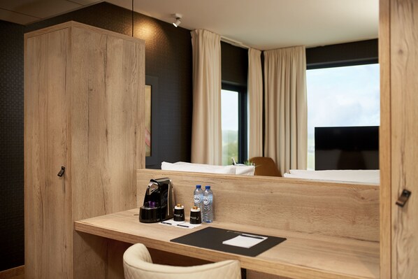 A hotel room with a wooden desk, a chair, a minibar, a bed, and a large window with curtains.