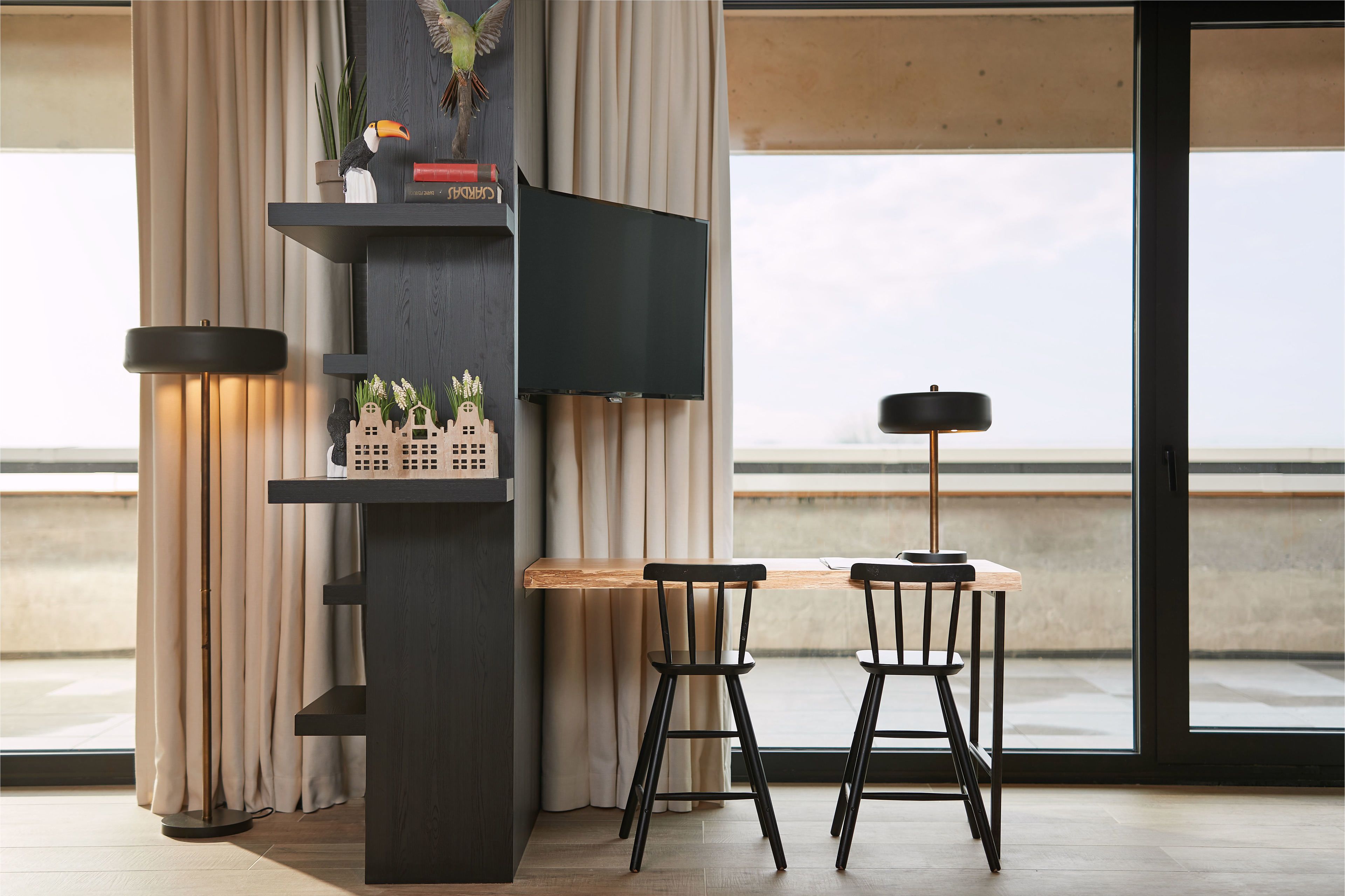 Un espace repas moderne avec une table en bois et deux chaises noires, un téléviseur fixé à un mur sombre et un lampadaire.