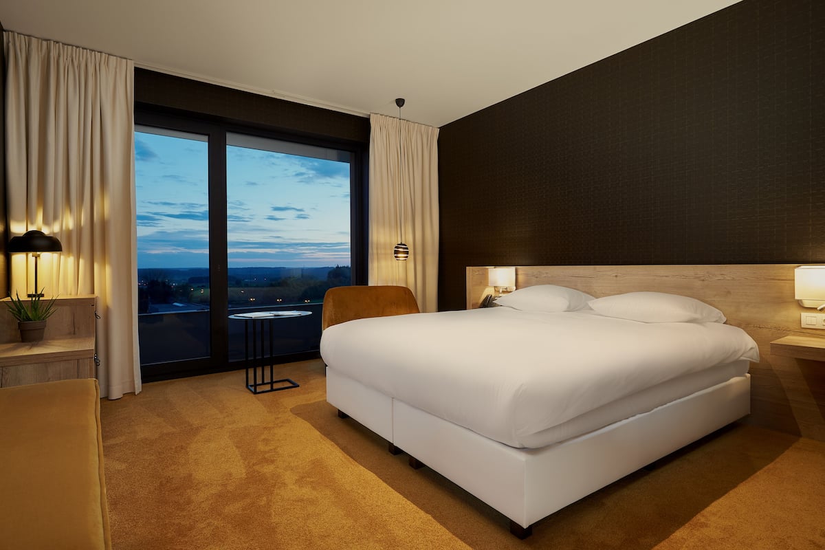 Habitación de hotel con una cama grande, una silla, una mesita y vista a la ciudad al anochecer.