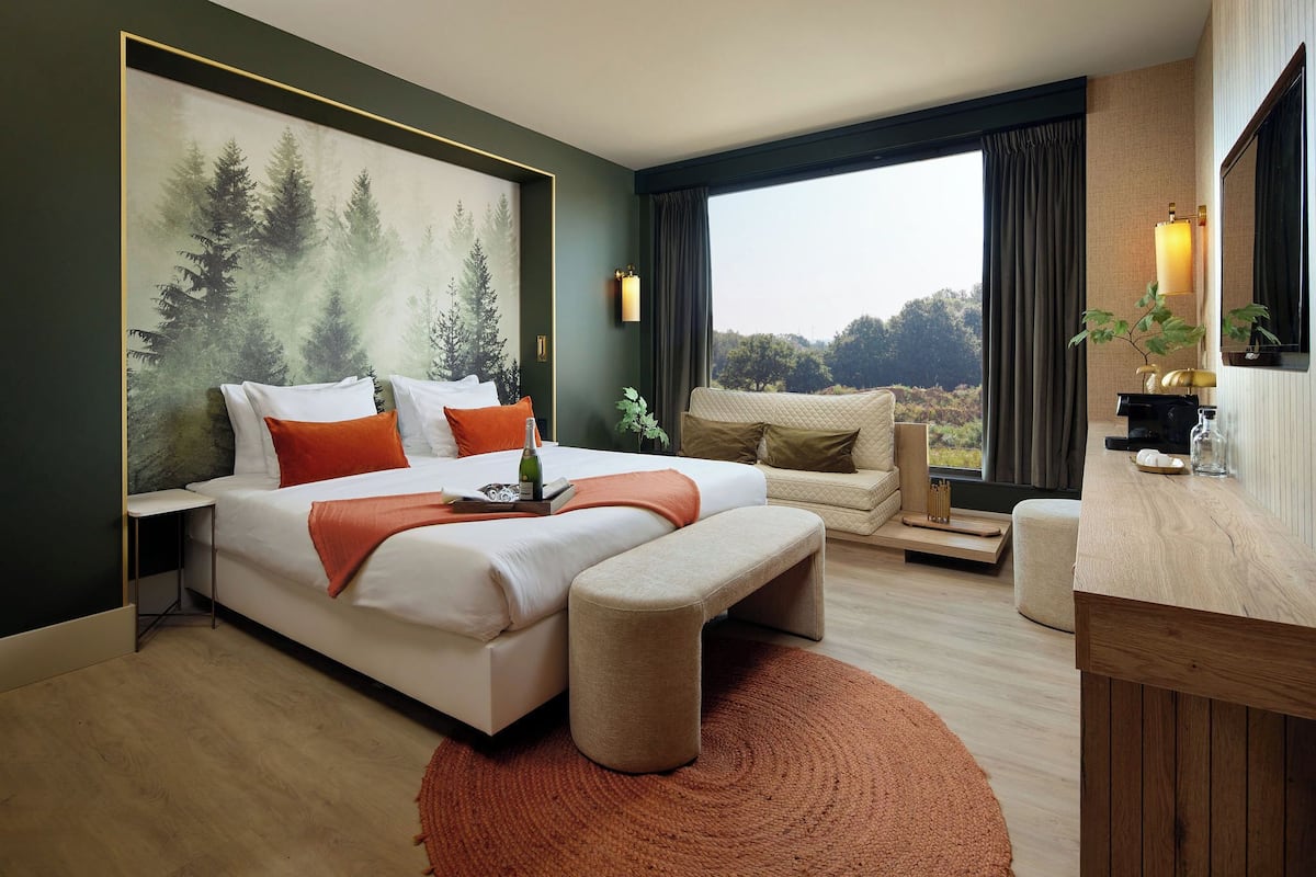 Una habitación de hotel moderna con una cama grande, un sofá, un escritorio de madera y un ventanal con vistas a árboles.