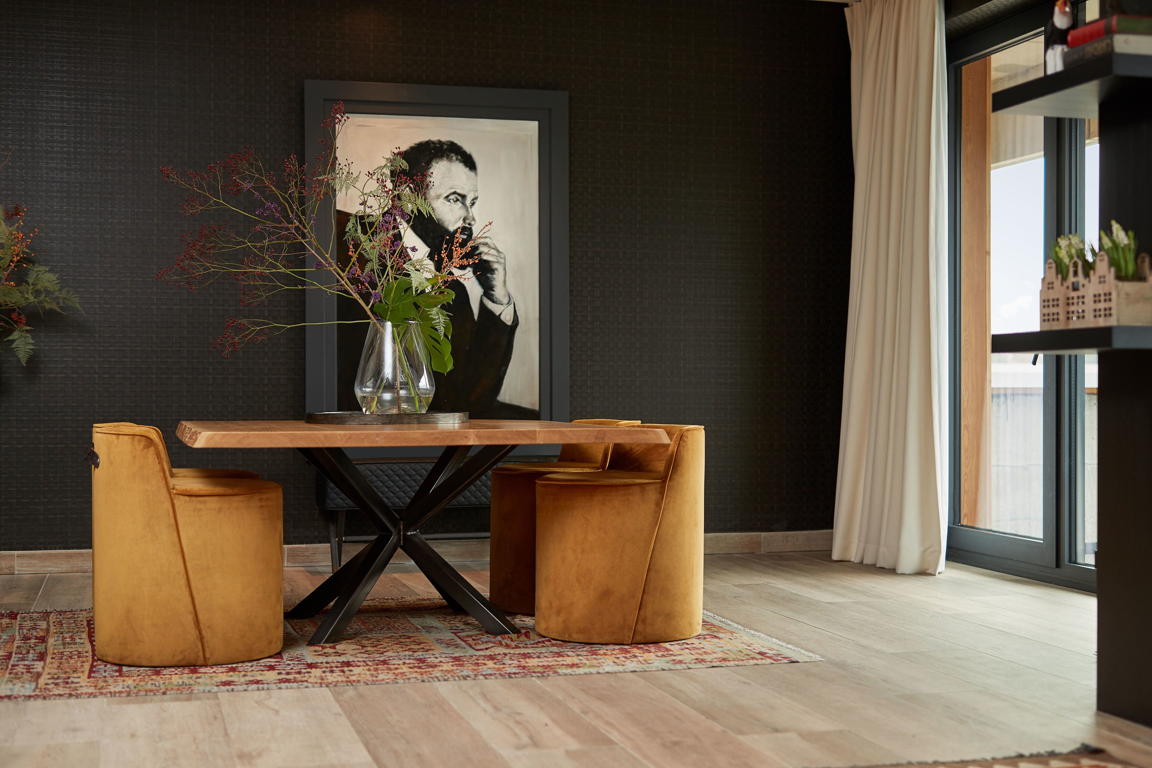 A dining area with a wooden table, two cushioned stools, a vase with flowers, and a framed portrait on the wall.