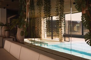 Un espace piscine intérieur moderne, avec des murs en pierre, de grandes fenêtres et des plantes d’intérieur.