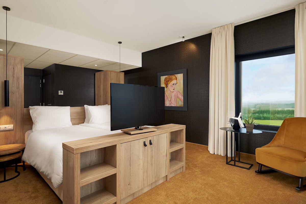 Habitación de hotel moderna con cama, televisión, consola de madera, sillón y un ventanal con vistas.
