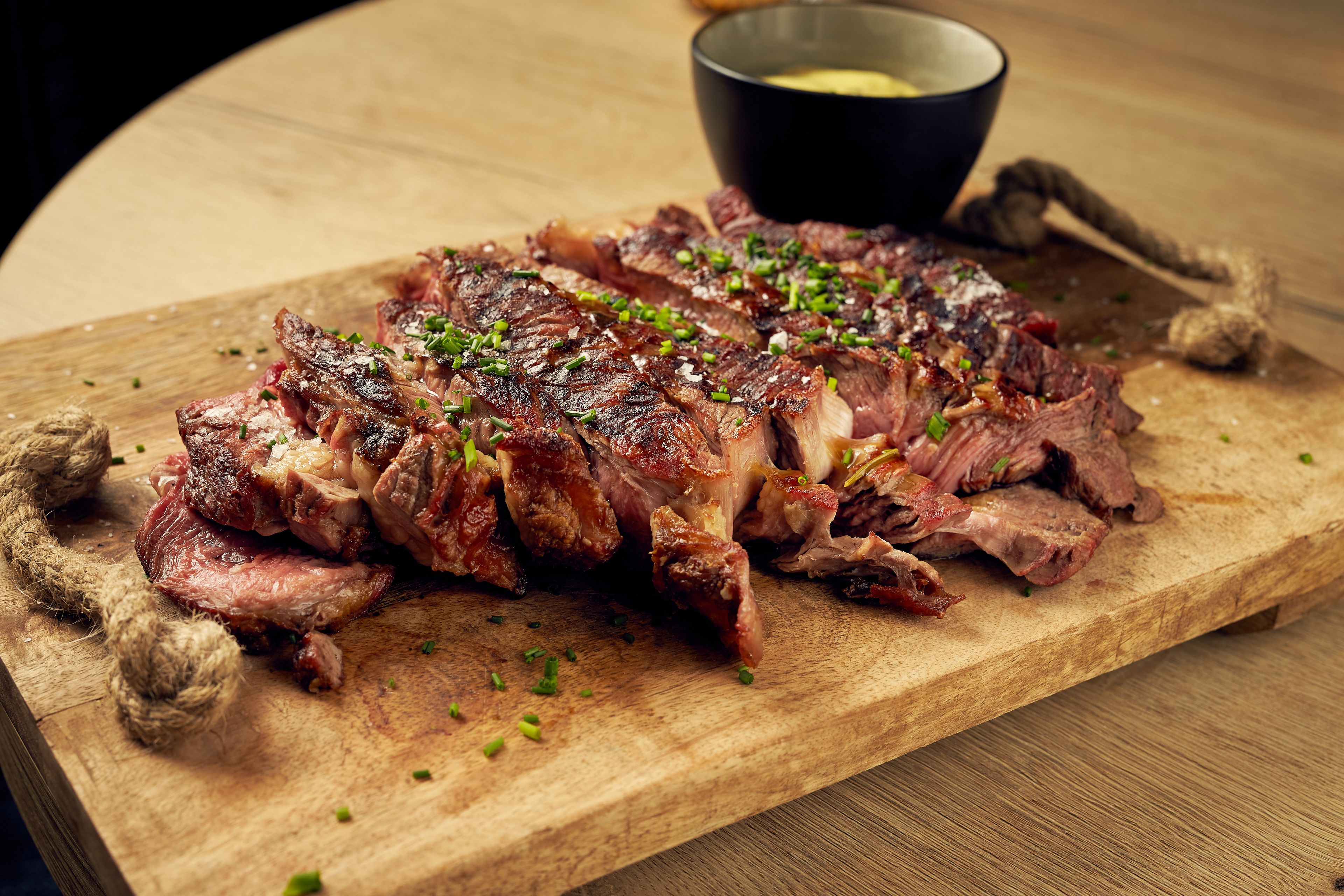 A wooden cutting board with sliced meat, garnished with herbs, and a bowl of sauce in the background.