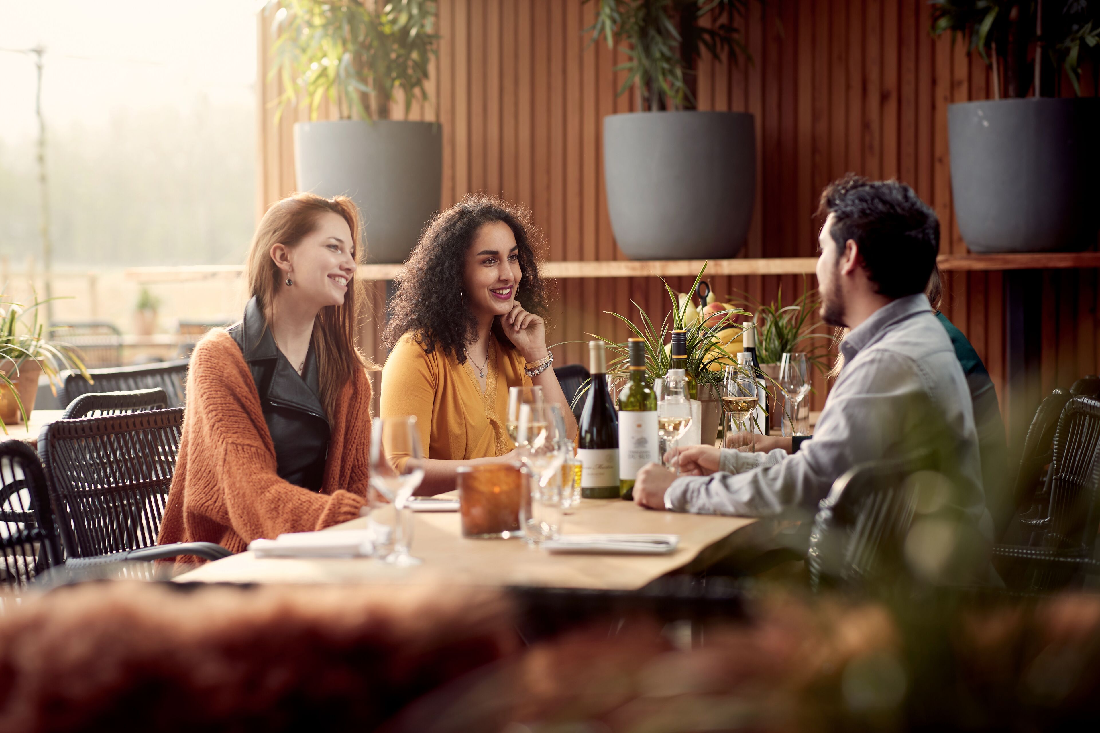 Un groupe de personnes est assis à une table à l’extérieur, entouré de plantes en pot et de bouteilles de vin.