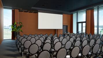 Un salón de conferencias con filas de sillas, una pantalla de proyección grande y una planta en maceta.
