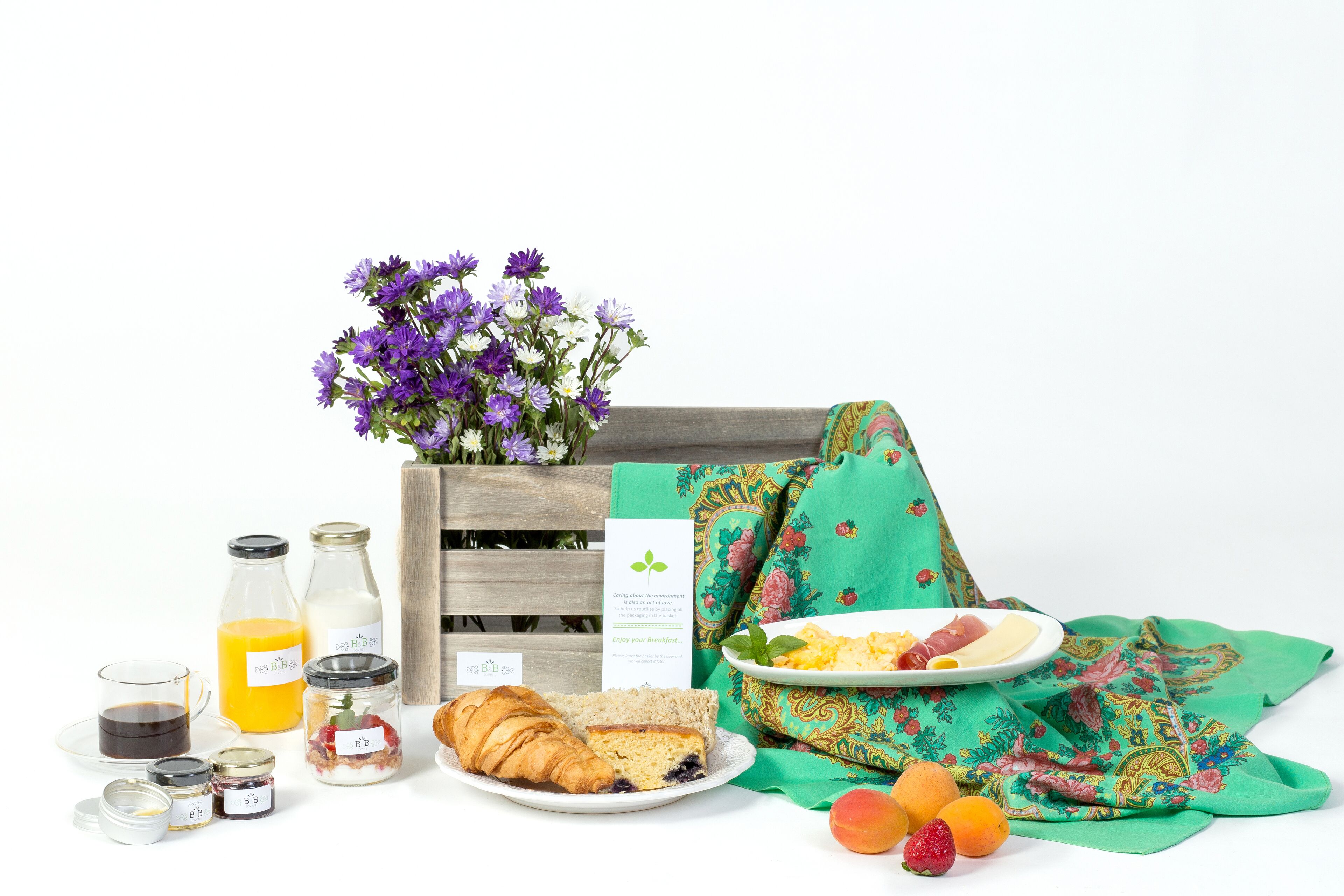 A breakfast setup with a wooden crate, a green cloth, a plate with eggs, toast, and fruit, and various breakfast items like juice, jam, and a croissant.