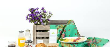 A breakfast setup with a wooden crate, a green cloth, a plate with eggs, toast, and fruit, and various breakfast items like juice, jam, and a croissant.