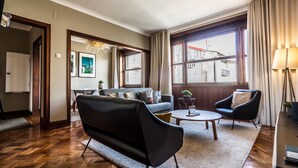 A modern living room with a sofa, armchair, and coffee table. There is a large window with curtains, a lamp, and a view of buildings outside. - YOUROPO - Santa Catarina (Porto)