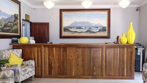A reception area with a wooden desk, two chairs, a potted plant, and a large framed painting of a coastal landscape.