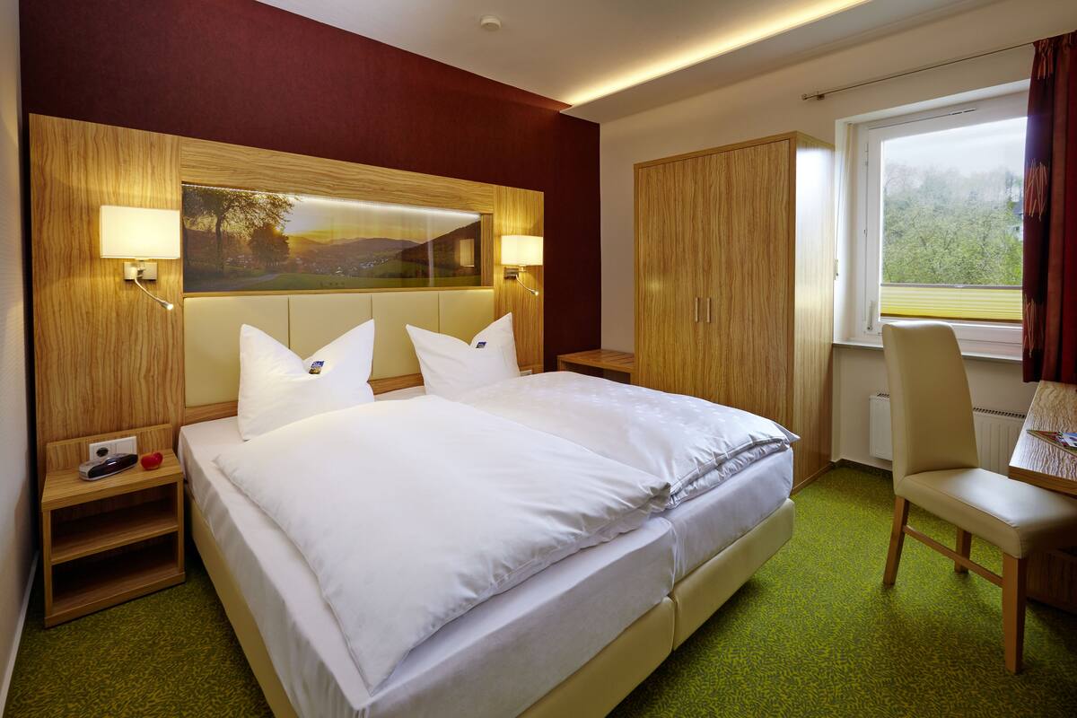 Quarto de hotel com uma cama grande, duas mesas de cabeceira, uma escrivaninha e uma janela com vista para árvores.