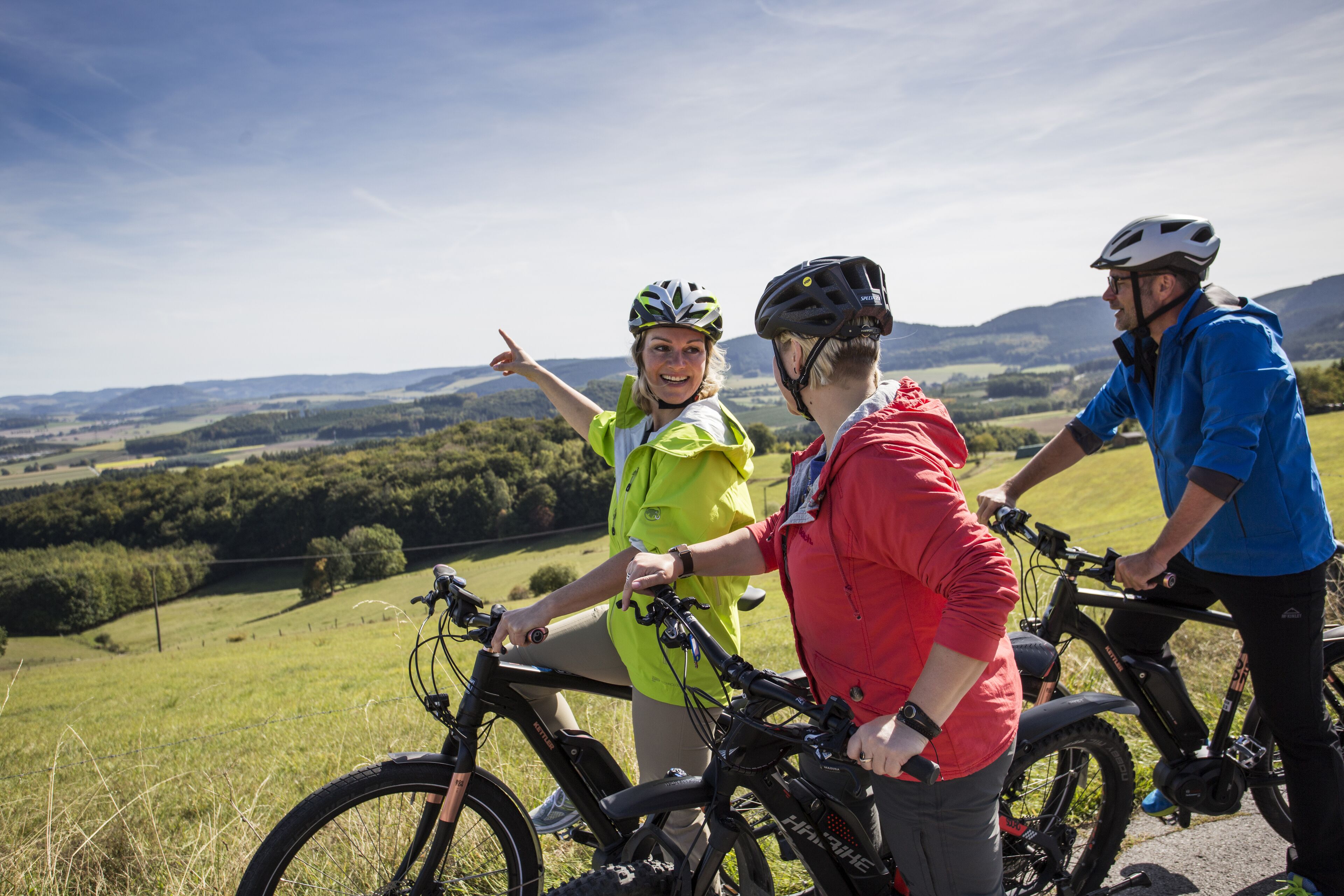 Three people on a mountain bike trail with a scenic view of rolling hills and a clear sky.