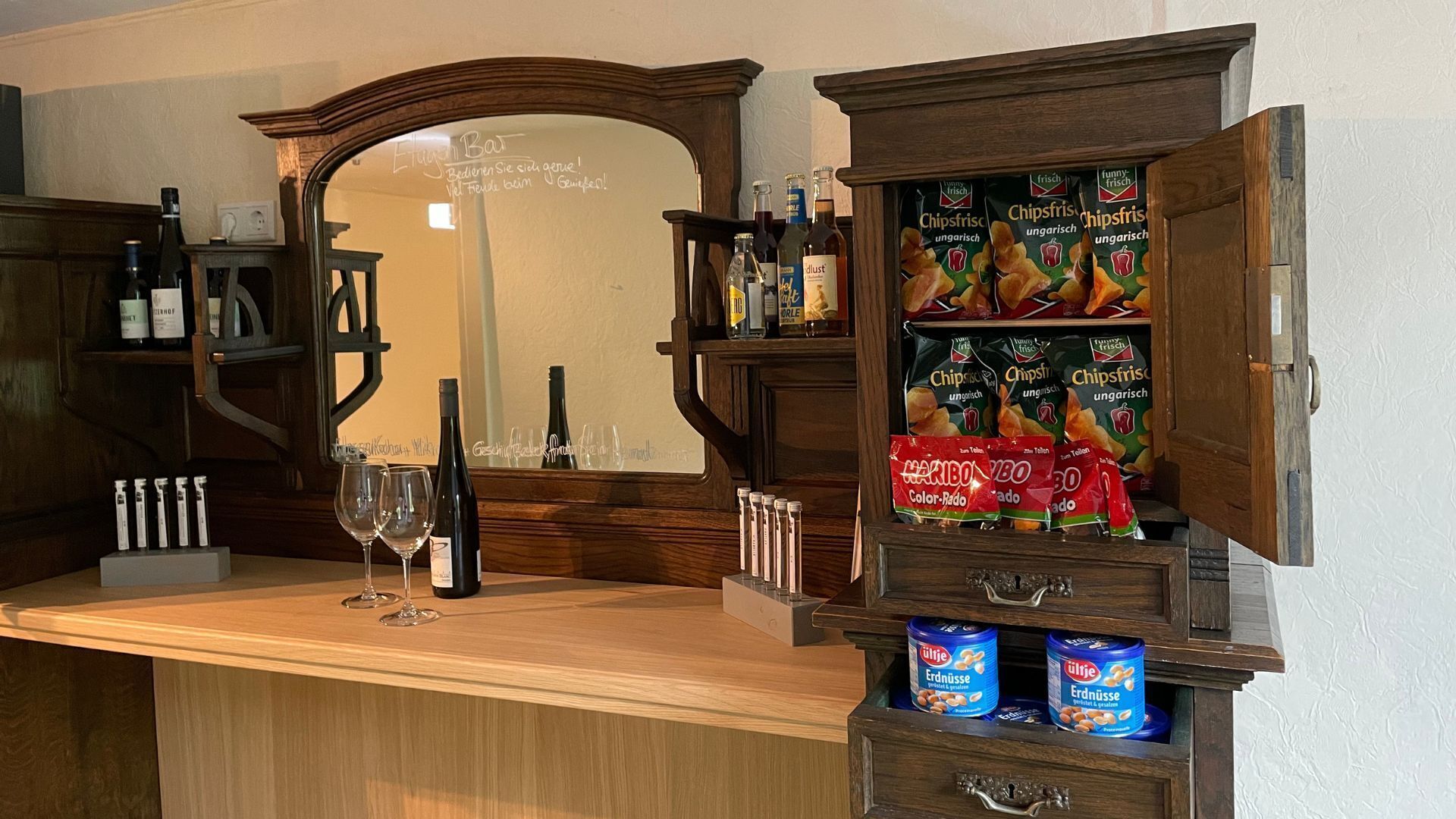 A bar area with a mirror, wine glasses, bottles, snacks, and a cabinet with drawers.
