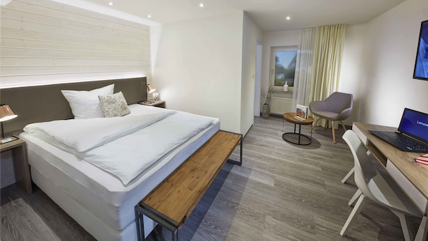Een moderne hotelkamer met een bed, nachtkastje, lamp, bureau met een laptop, stoel en uitzicht op het buiten.