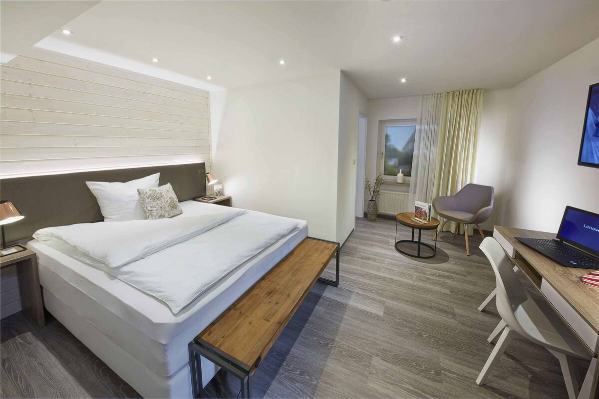Quarto de hotel moderno com cama, mesa de cabeceira, luminária, escrivaninha com um laptop, cadeira e vista para o exterior.