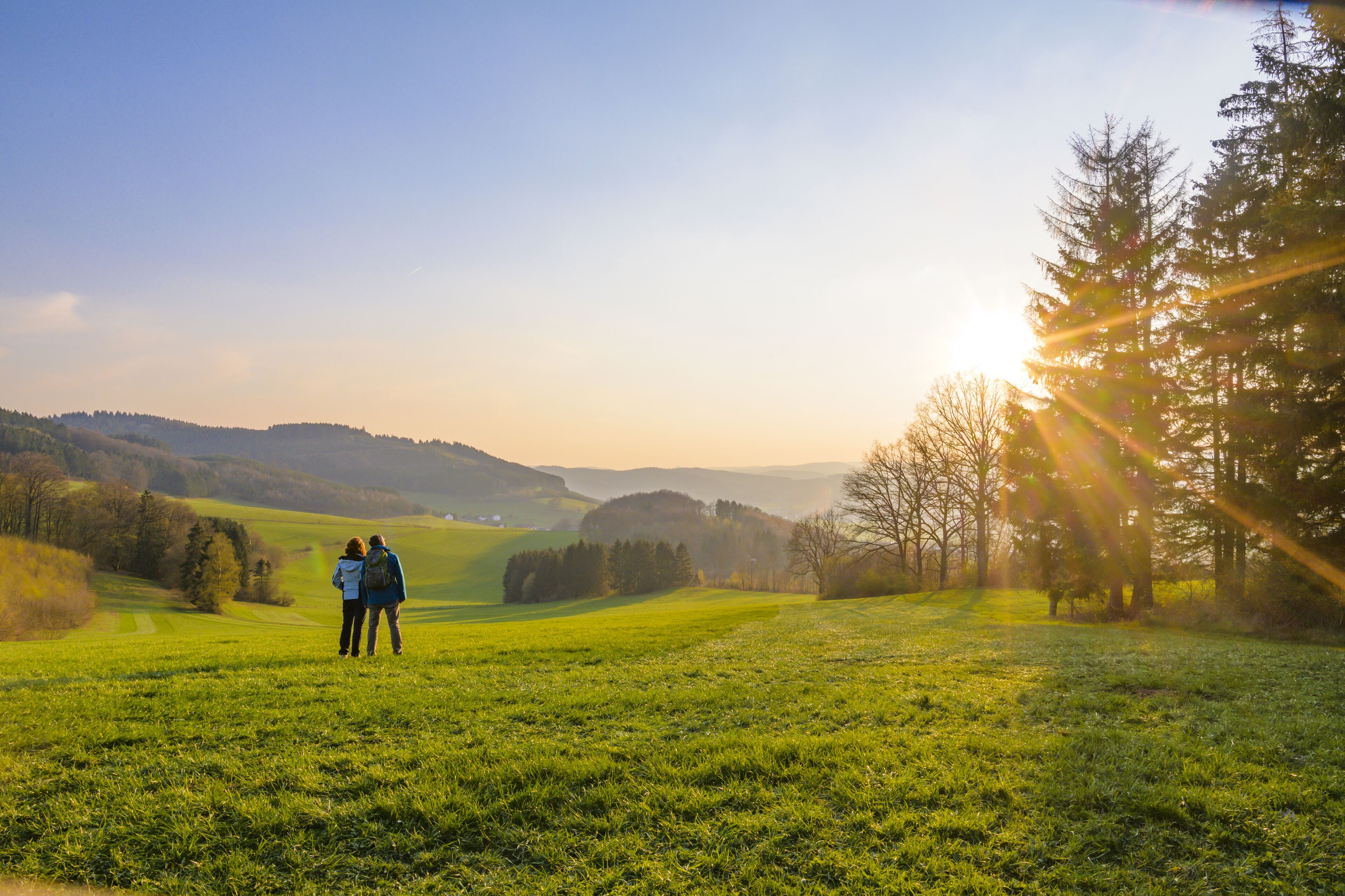 Ein Paar steht auf einer Wiese mit Blick auf sanfte Hügel und einen Sonnenuntergang im Hintergrund.