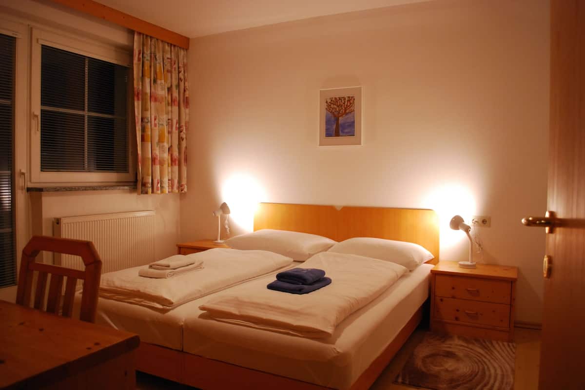 Un dormitorio con una cama doble, dos mesitas de noche con lámparas, un cabecero de madera, un radiador y una ventana con persianas.