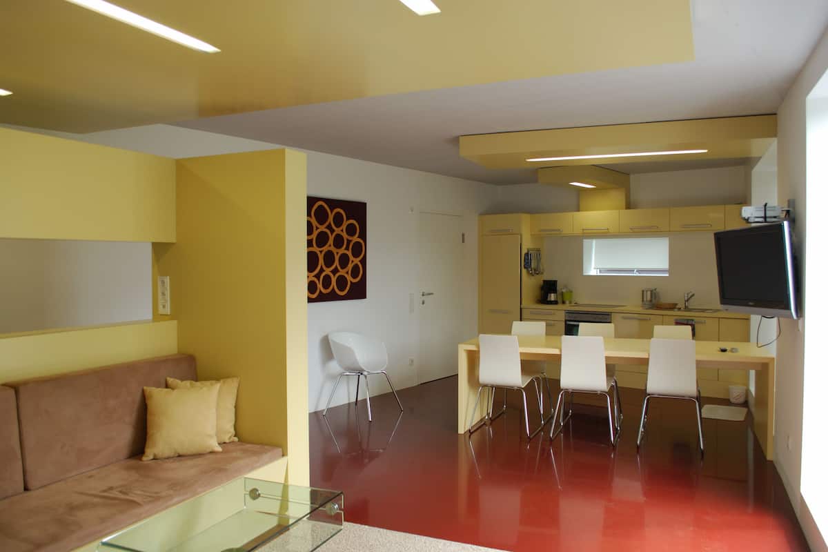 Una sala moderna con sofá y mesa de centro de vidrio, un comedor con sillas y mesa, y una cocina con armarios y televisor.