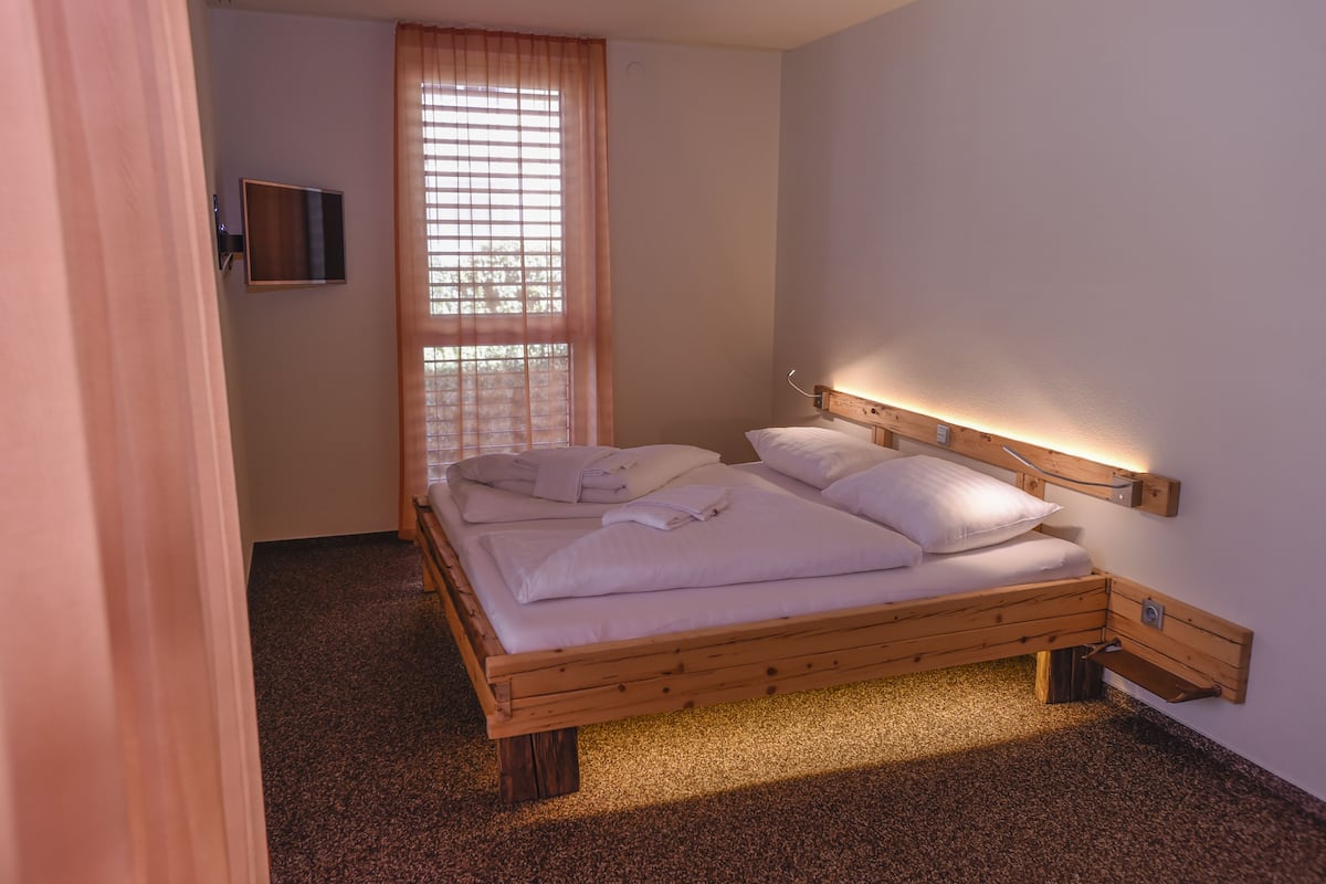 Una cama individual con cabecera y piecera de madera, un televisor montado en la pared y una ventana con persianas.
