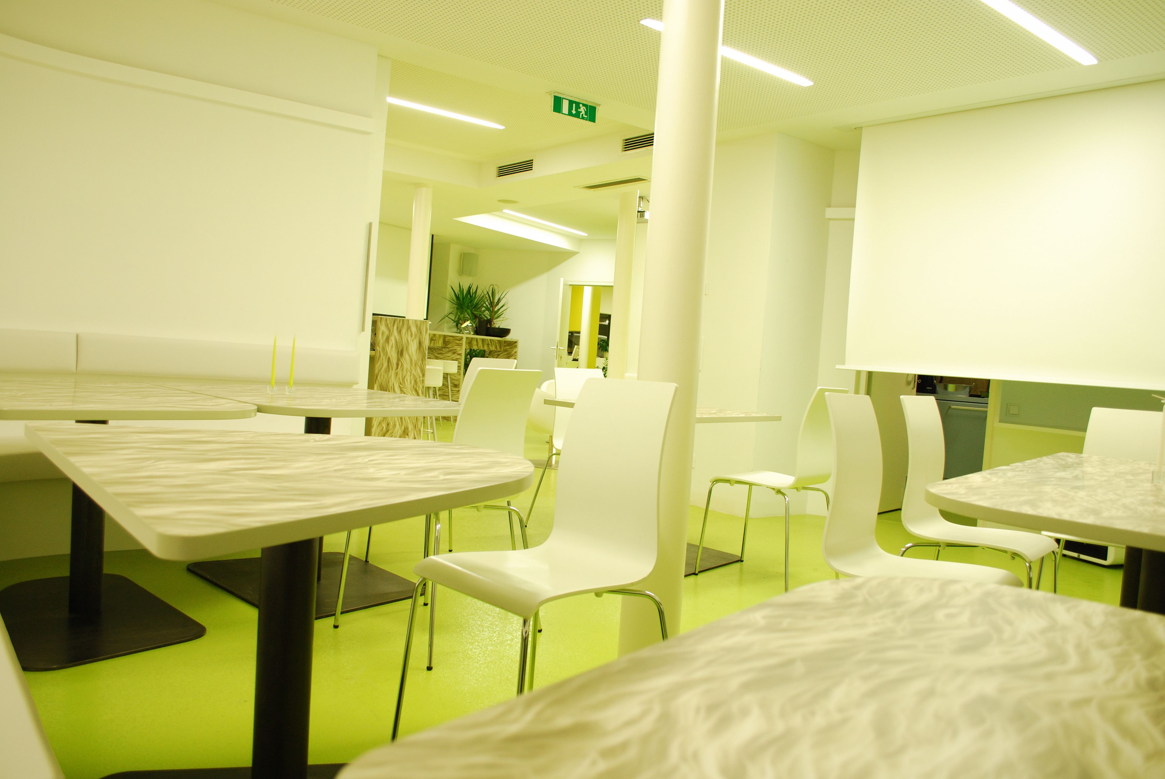 Uma sala de jantar moderna com mesas e cadeiras brancas, piso verde e uma saída identificada.