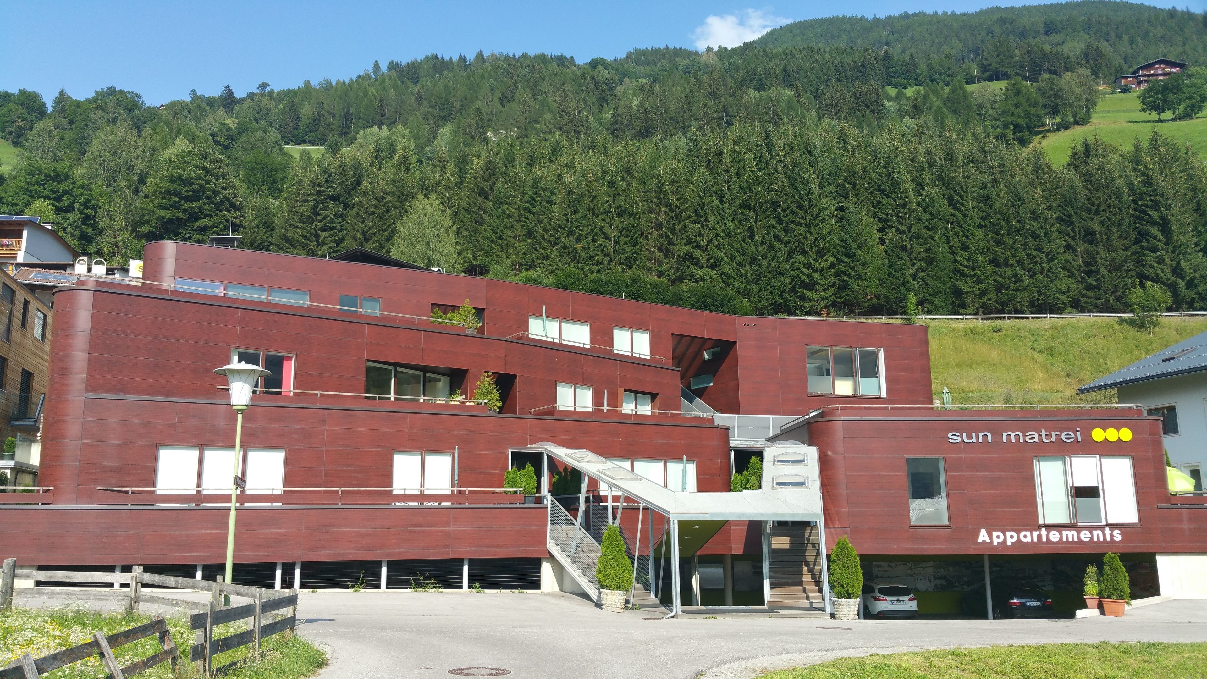 A modern red building with a staircase leading to a covered entrance, surrounded by greenery and a hilly landscape.
