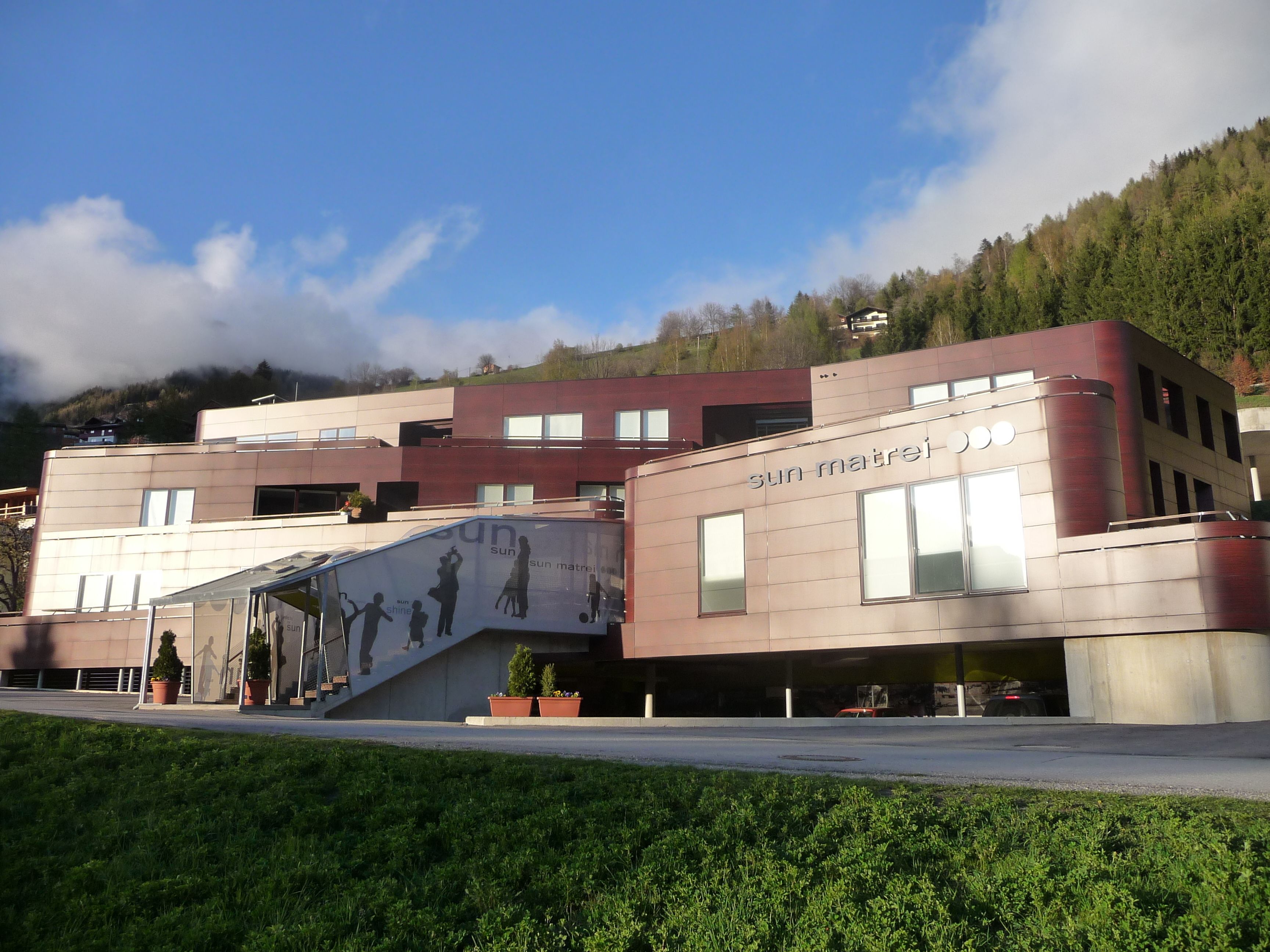 A modern building with a ramp leading to the entrance, surrounded by greenery and a clear sky.