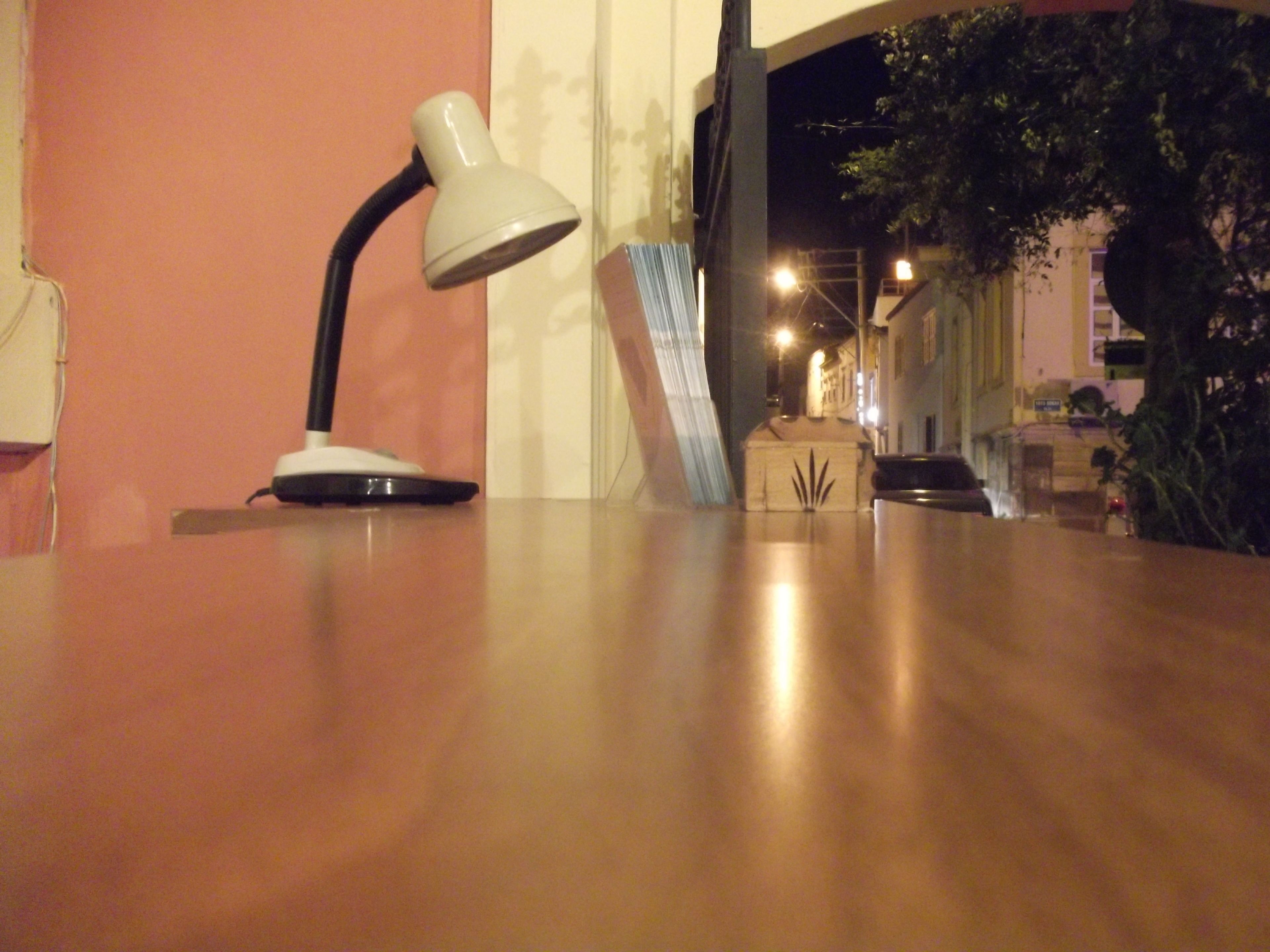 Un escritorio con una lámpara, un pequeño cofre de madera y una ventana que da a una calle y edificios de noche.
