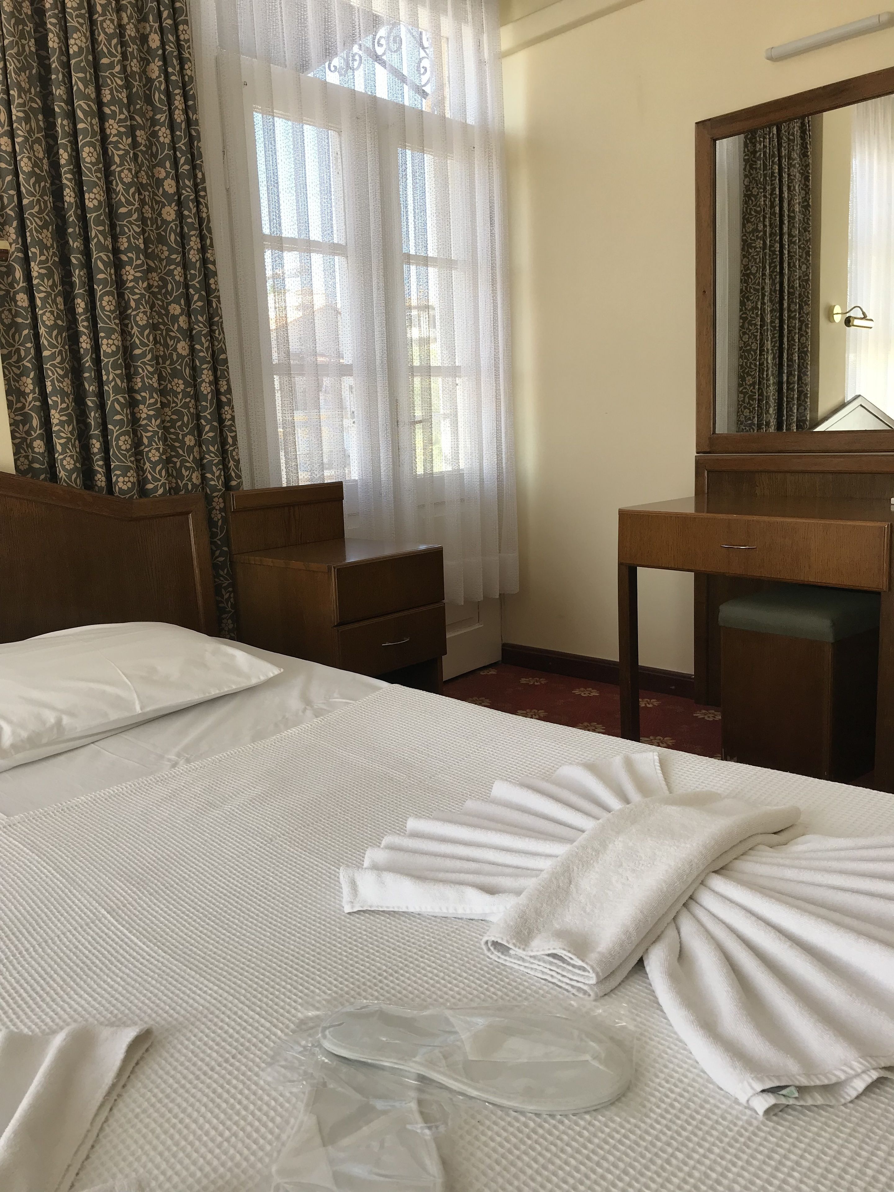 Hotellihuone, jossa on sänky, puinen yöpöytä, peili ja ikkuna, jossa on läpikuultavat verhot.