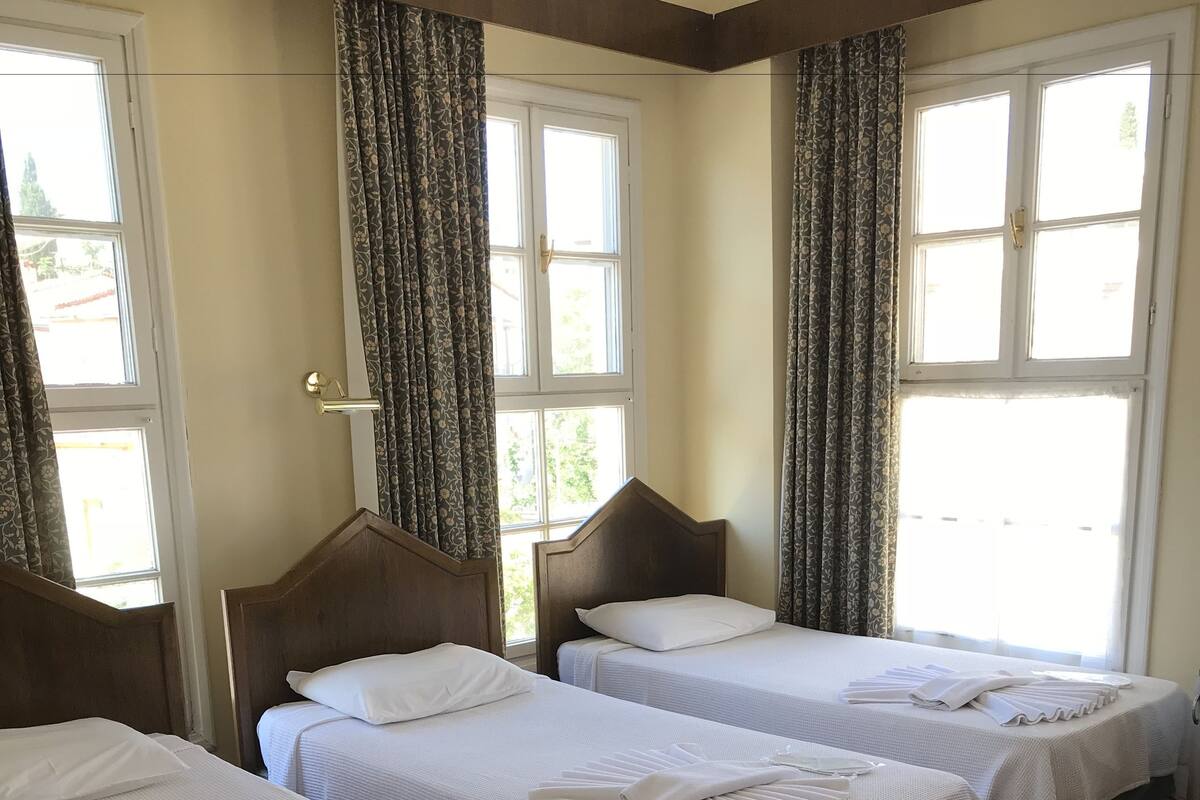A hotel room with three single beds, each with a neatly folded towel and a white bedspread. The room has a red floral carpet and large windows with curtains.