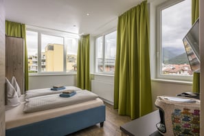 A hotel room with two beds, a desk with a computer, and a view of buildings and mountains through large windows. - Backstage Kufstein (Kufstein)