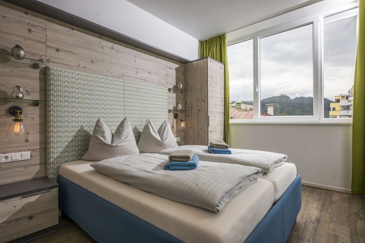 Ein modernes Schlafzimmer mit zwei Betten, einem Holz-Kopfteil, einem Fenster mit Blick auf Gebäude und einem grünen Vorhang.