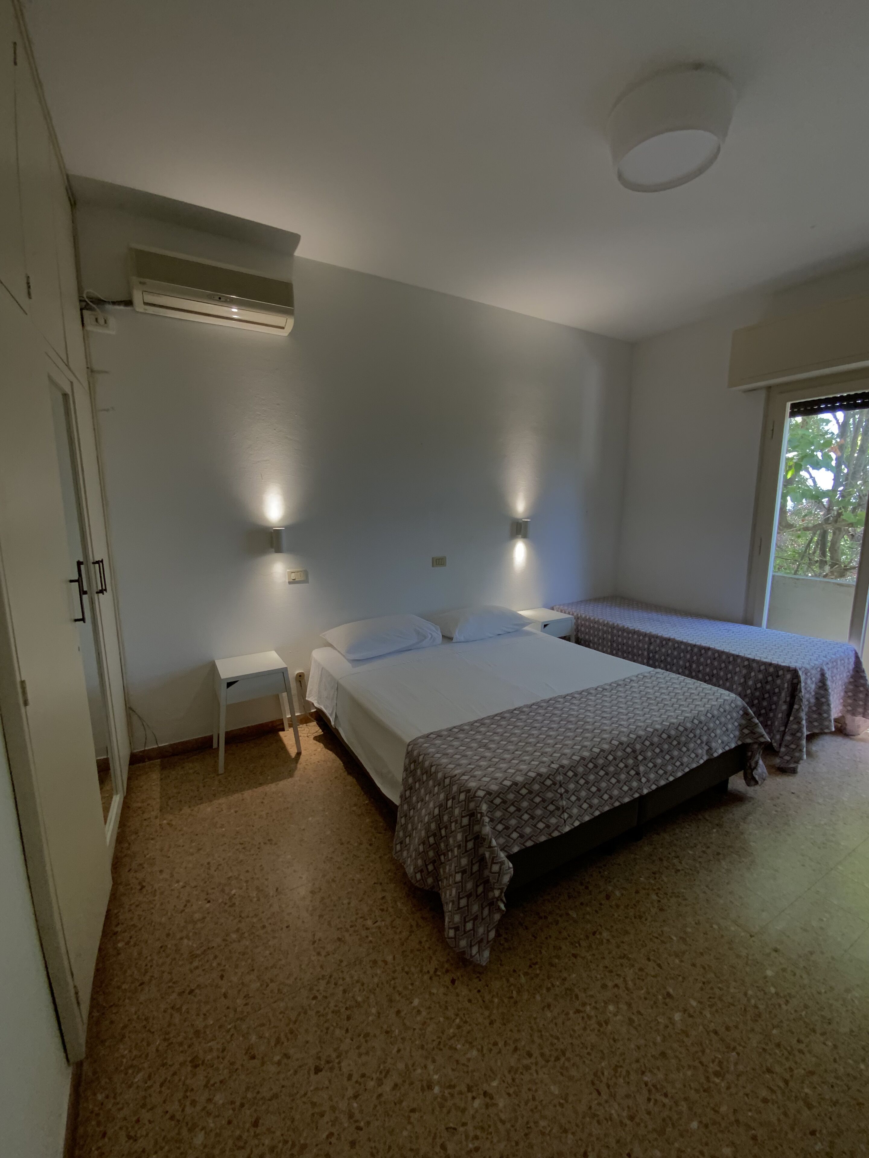Un dormitorio con dos camas, una mesita de noche pequeña, aire acondicionado instalado en la pared y una ventana con vista a árboles.