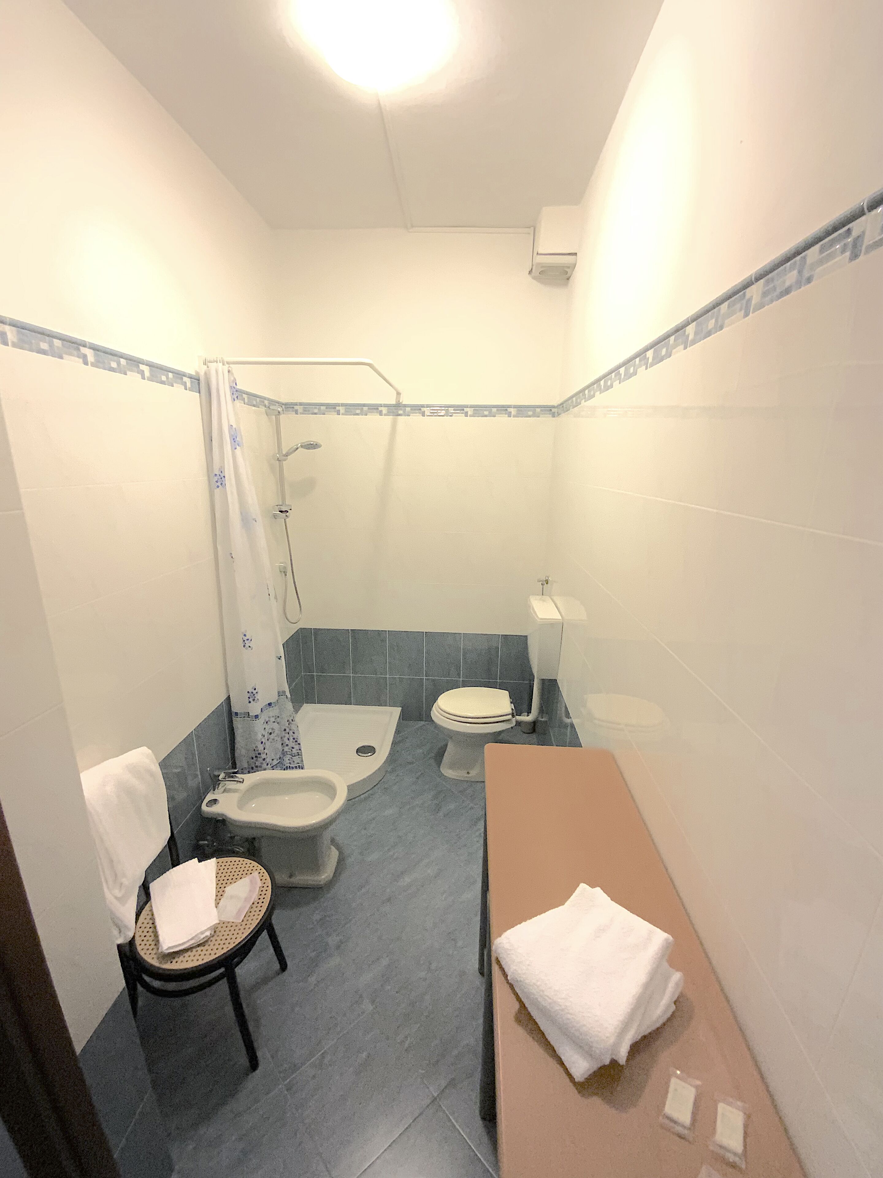 A small bathroom with a shower, two toilets, a sink, and a chair.