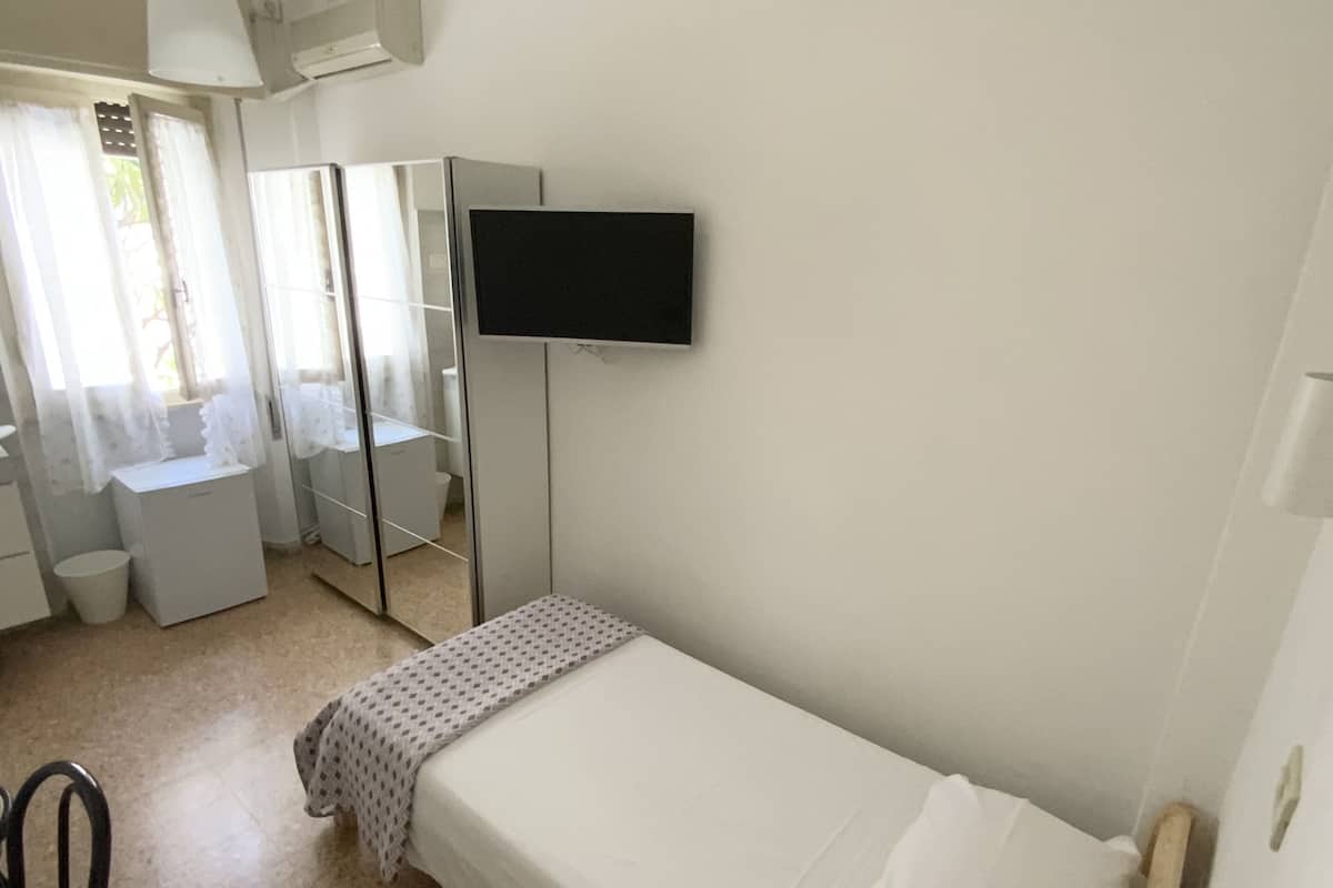 A small, clean room with a single bed, a TV mounted on the wall, and a window with sheer curtains.