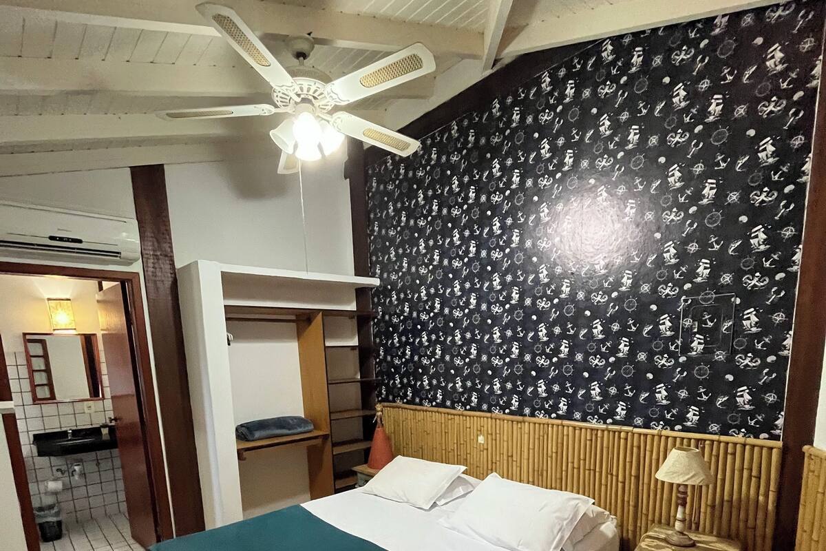 Hotelkamer met een bed, een plafondventilator, een muur met een nautisch patroon, een tafeltje met een lamp en een badkamer die door een open deur zichtbaar is.