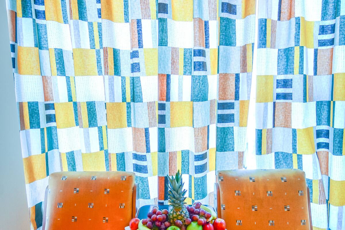 Une pièce avec un rideau à carreaux, deux fauteuils orange, une petite table sur laquelle se trouvent des fruits et un ananas en guise de décoration centrale.