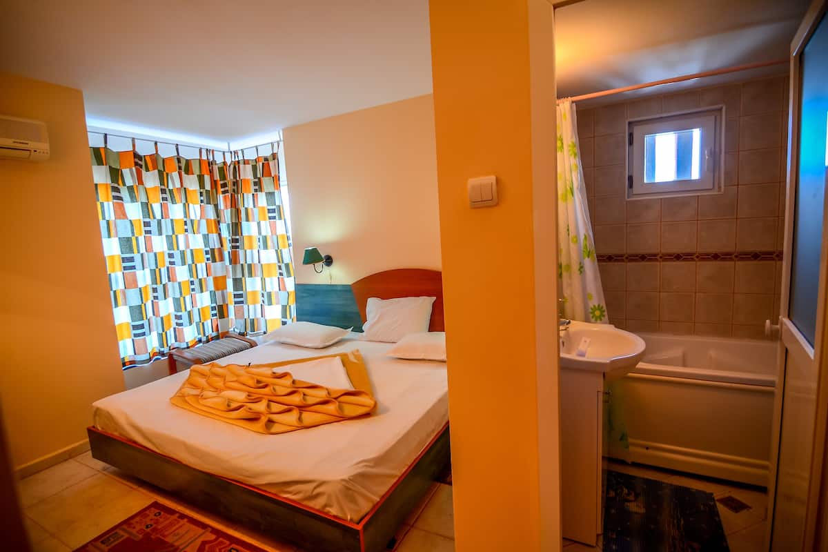 Une chambre d’hôtel avec un lit, une fenêtre ornée d’un rideau coloré, une salle de bain avec une baignoire et une petite fenêtre située au-dessus de la baignoire.