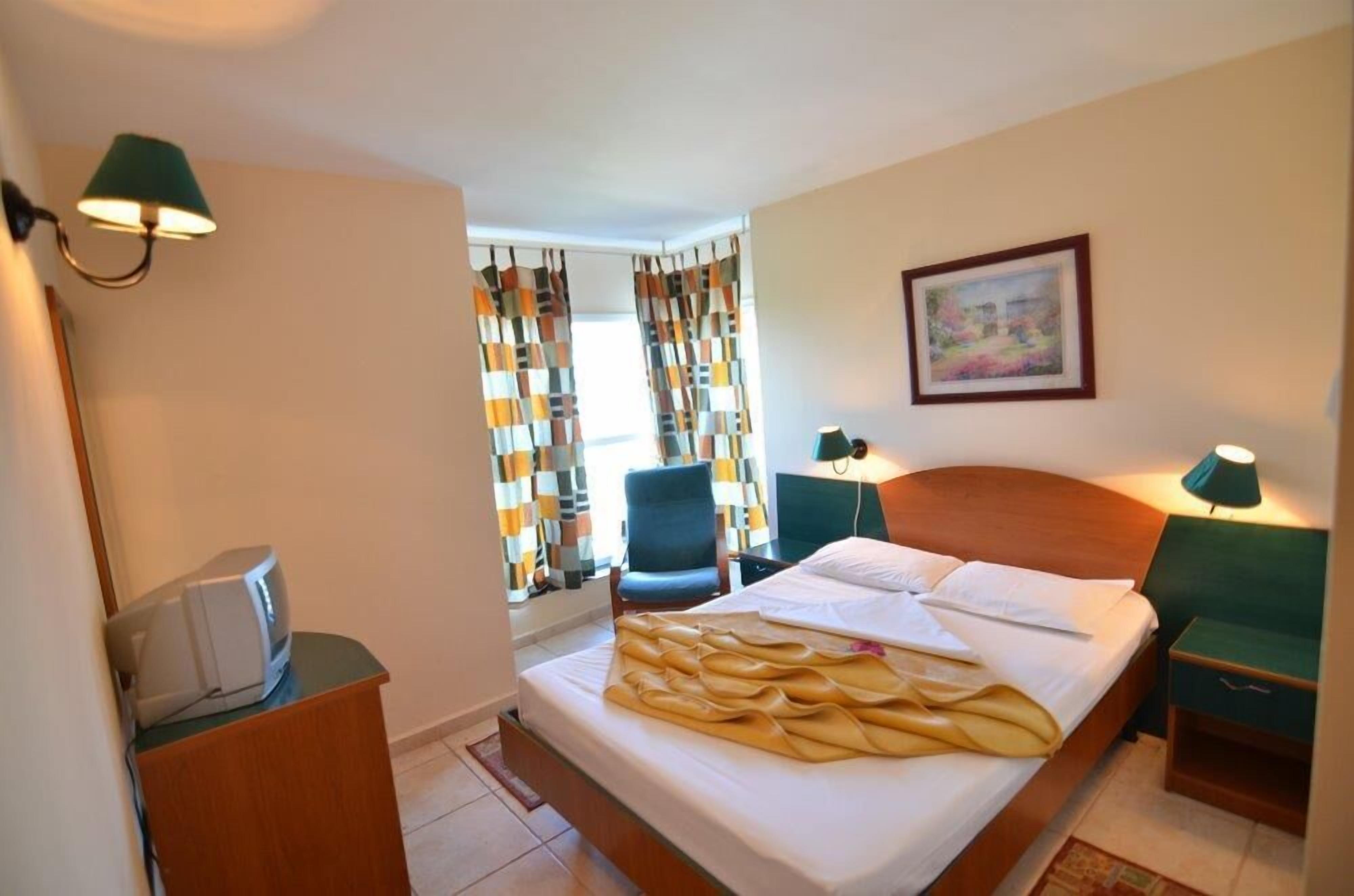 A hotel room with a bed, a chair, a desk with a lamp, and a painting on the wall.