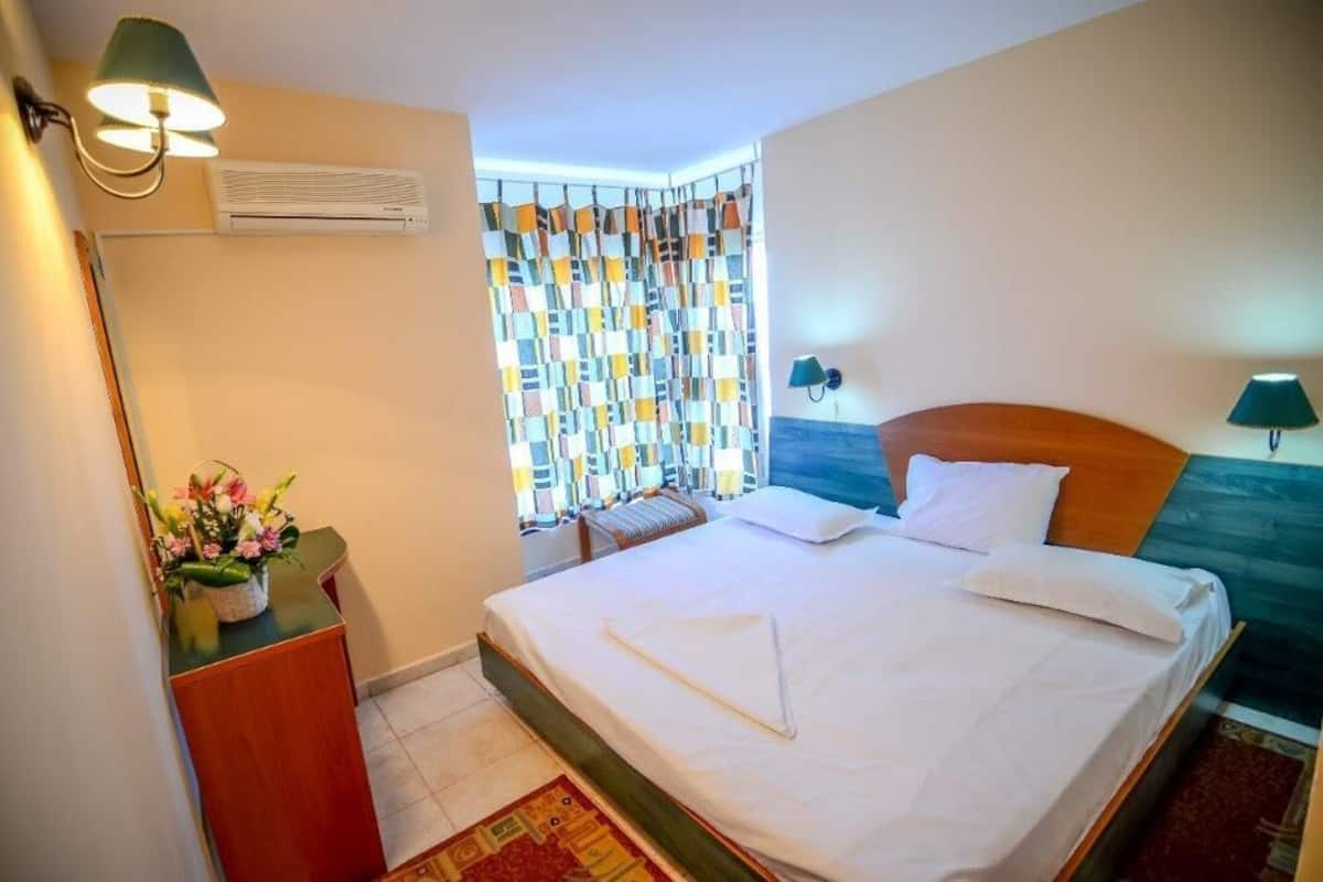 Une chambre d’hôtel comprenant un lit, des lampes de chevet, une fenêtre avec des rideaux, un appareil de climatisation, un bureau avec une lampe et un arrangement floral.