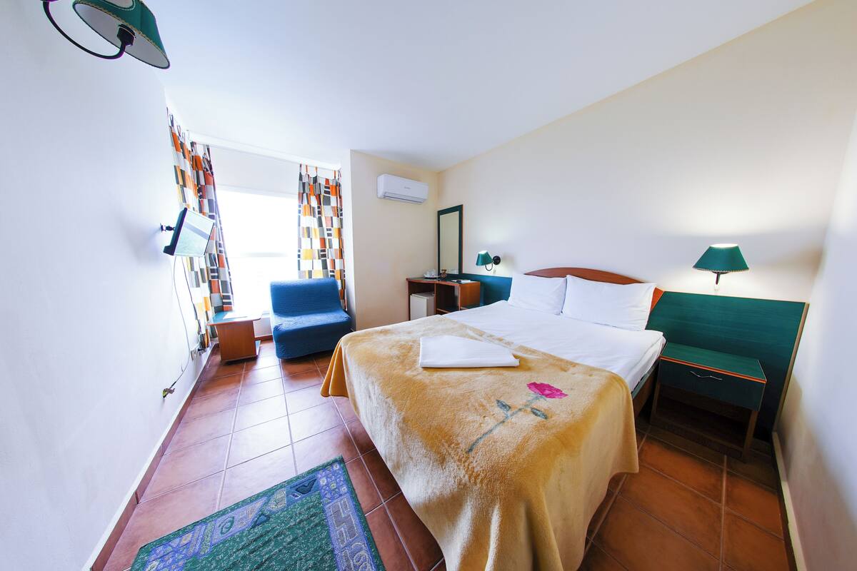 Une chambre d’hôtel comprenant un lit, un bureau, une chaise, une table de chevet, une lampe, un miroir et une fenêtre avec des rideaux.