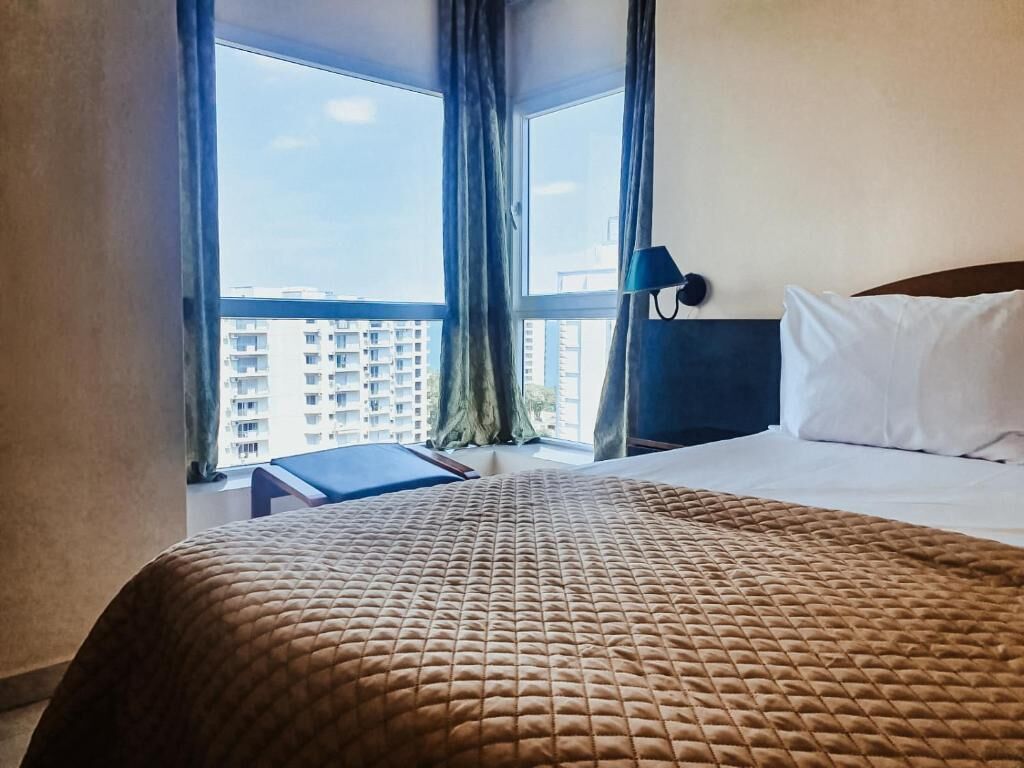 A hotel room with a bed, a desk, and a view of buildings through large windows.