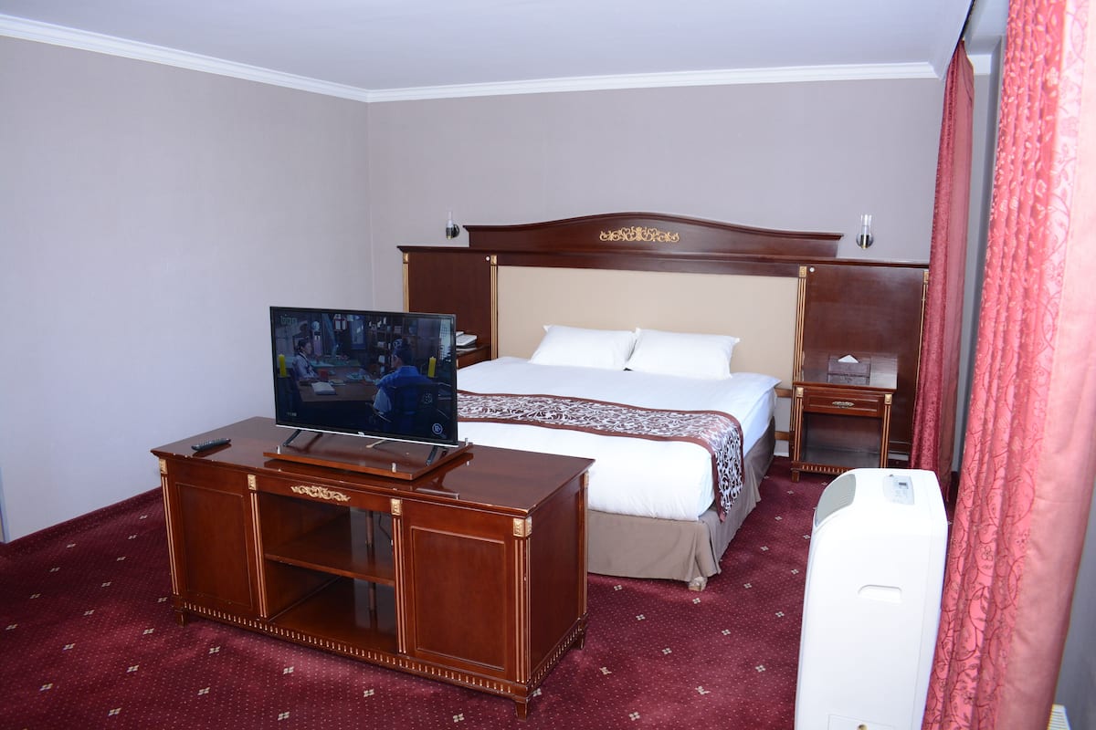 Hotellihuone, jossa on sänky, puupöydällä oleva televisio, yöpöytä ja valkoinen ilmastointilaite.