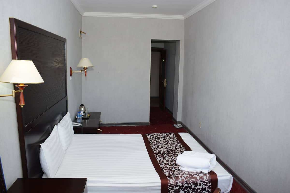Hotellihuone, jossa on suuri sänky, yöpöydällä olevat lamput, yöpöytä ja punaisella matolla päällystetty käytävä.