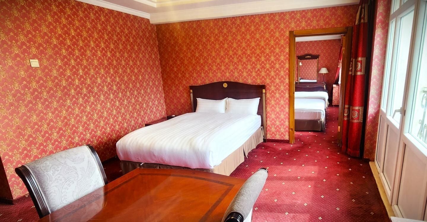 A hotel room with a large bed, a wooden table, and a chair. There is a mirror on the wall and a window with curtains.