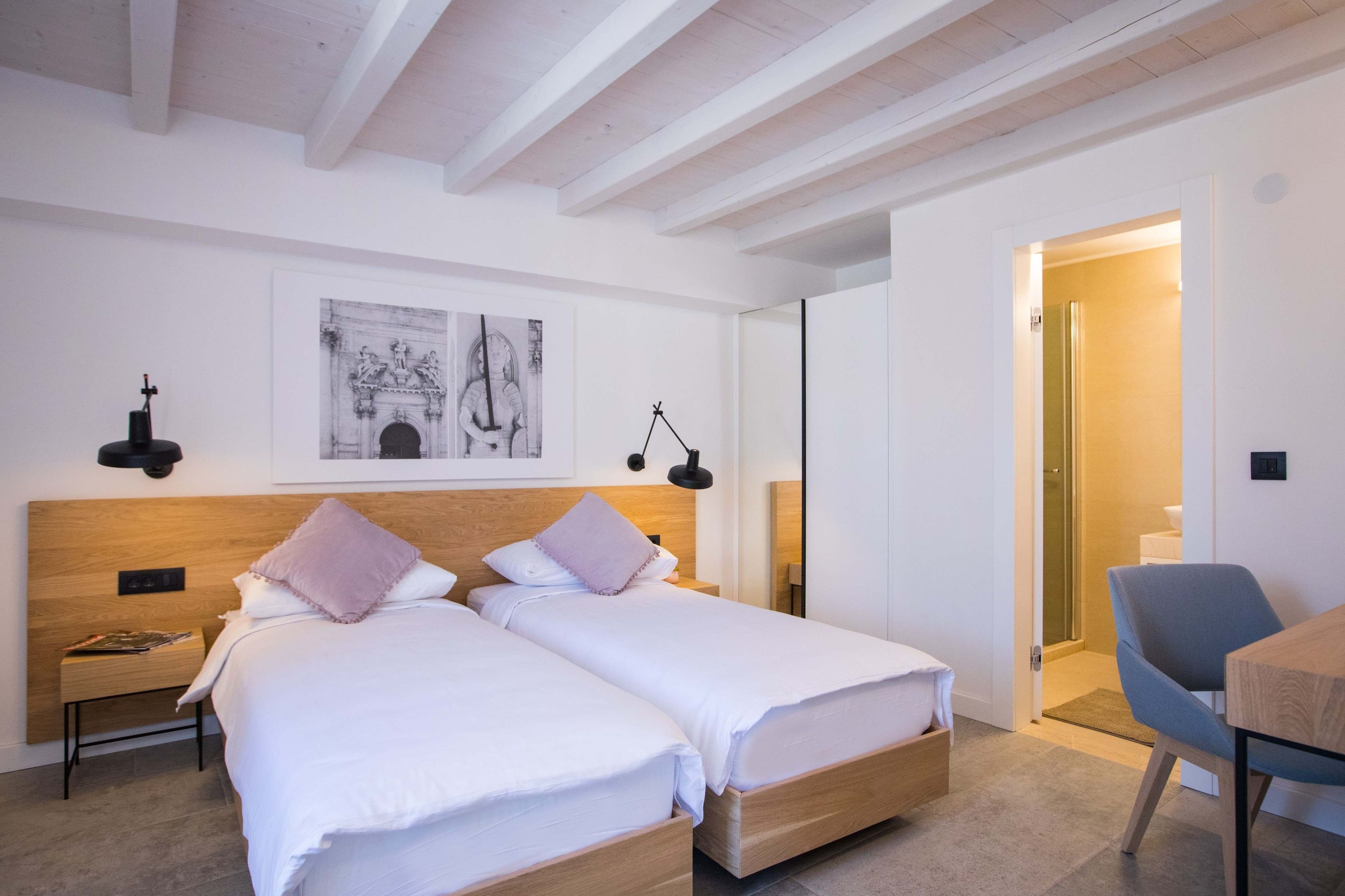A modern bedroom with two single beds, a wooden headboard, a nightstand, a desk, and a chair.